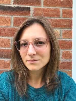 Portrait de Rachel, femme aux cheveux bruns mi-longs portant des lunettes roses et un chandail turquoise, devant un mur de briques