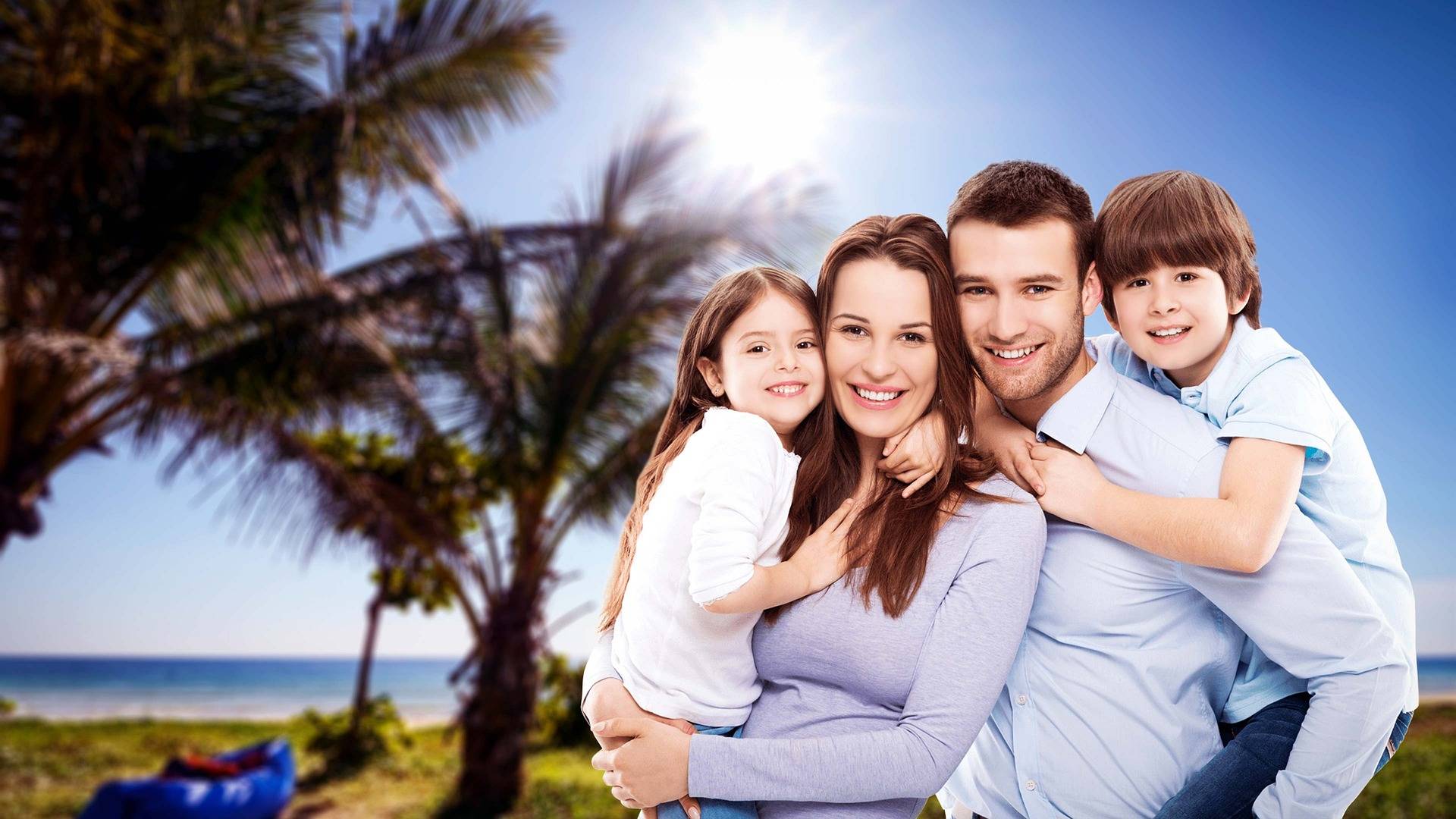 Une famille de quatre personnes sourit face à la caméra sur une plage ensoleillée. Les deux parents sont au centre, entourés de leurs deux enfants qui les enlacent affectueusement. À l'arrière-plan, on aperçoit des palmiers, le ciel bleu, le soleil et la mer, créant une ambiance chaleureuse et estivale.
