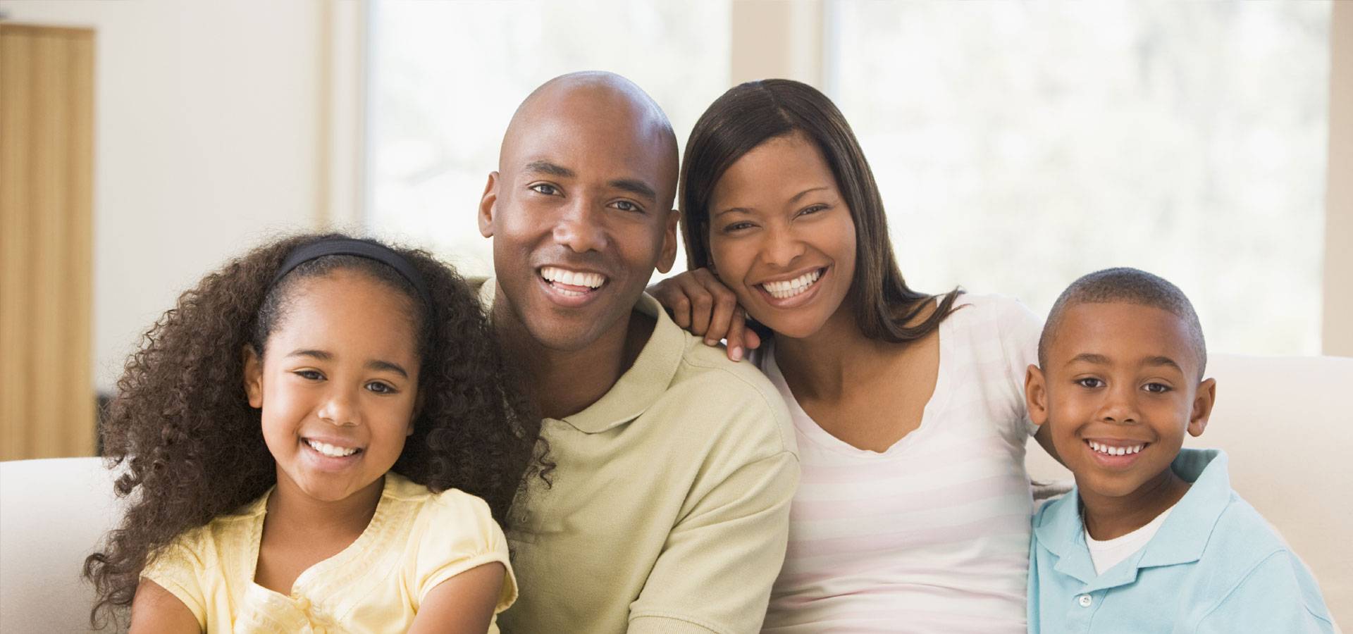 Une famille de quatre personnes sourit à la caméra dans un intérieur lumineux. Les deux parents sont assis au centre, entourés de leurs deux enfants, une fille aux cheveux bouclés portant un haut jaune et un garçon portant un polo bleu clair. Tous regardent l'objectif avec un sourire, créant une ambiance chaleureuse et conviviale.