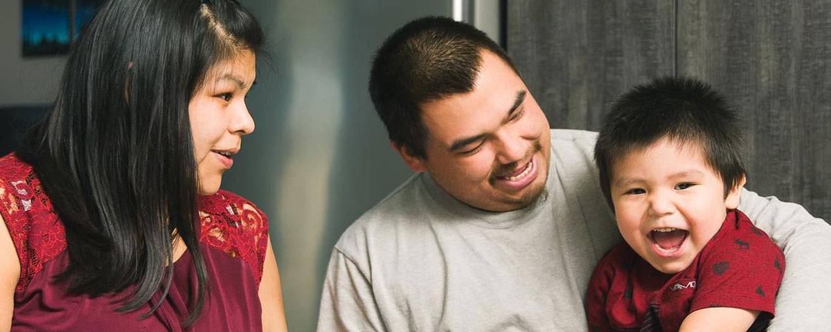 Une famille de trois personnes est réunie dans un intérieur. Un enfant sourit joyeusement en regardant la caméra, tandis que les deux adultes, assis de part et d'autre de lui, le regardent avec affection. L'ambiance est chaleureuse et met en valeur un moment de complicité familiale.